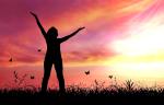 A woman standing in a field raising her arms to the sky with a sunset behind her