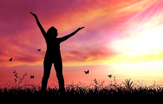 A woman standing in a field raising her arms to the sky with a sunset behind her